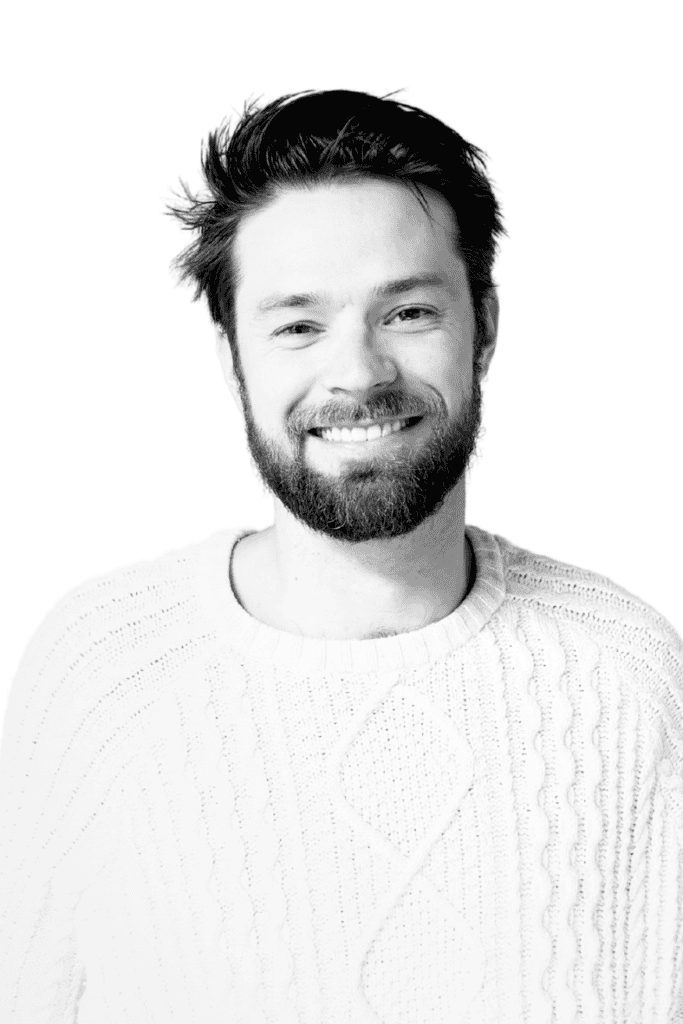 Smiling man with beard in a white sweater, professional headshot.
